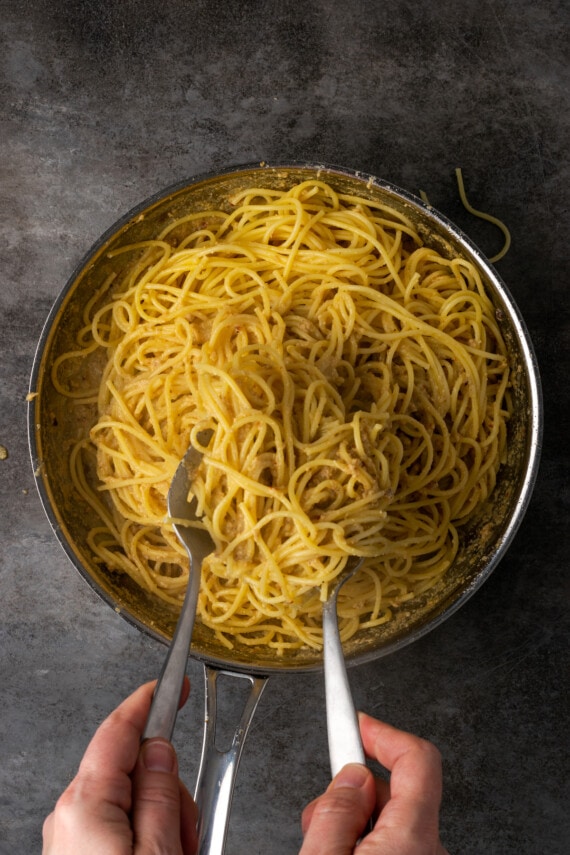 Una mano usando tenazas para mezclar pasta con salsa César en una sartén.