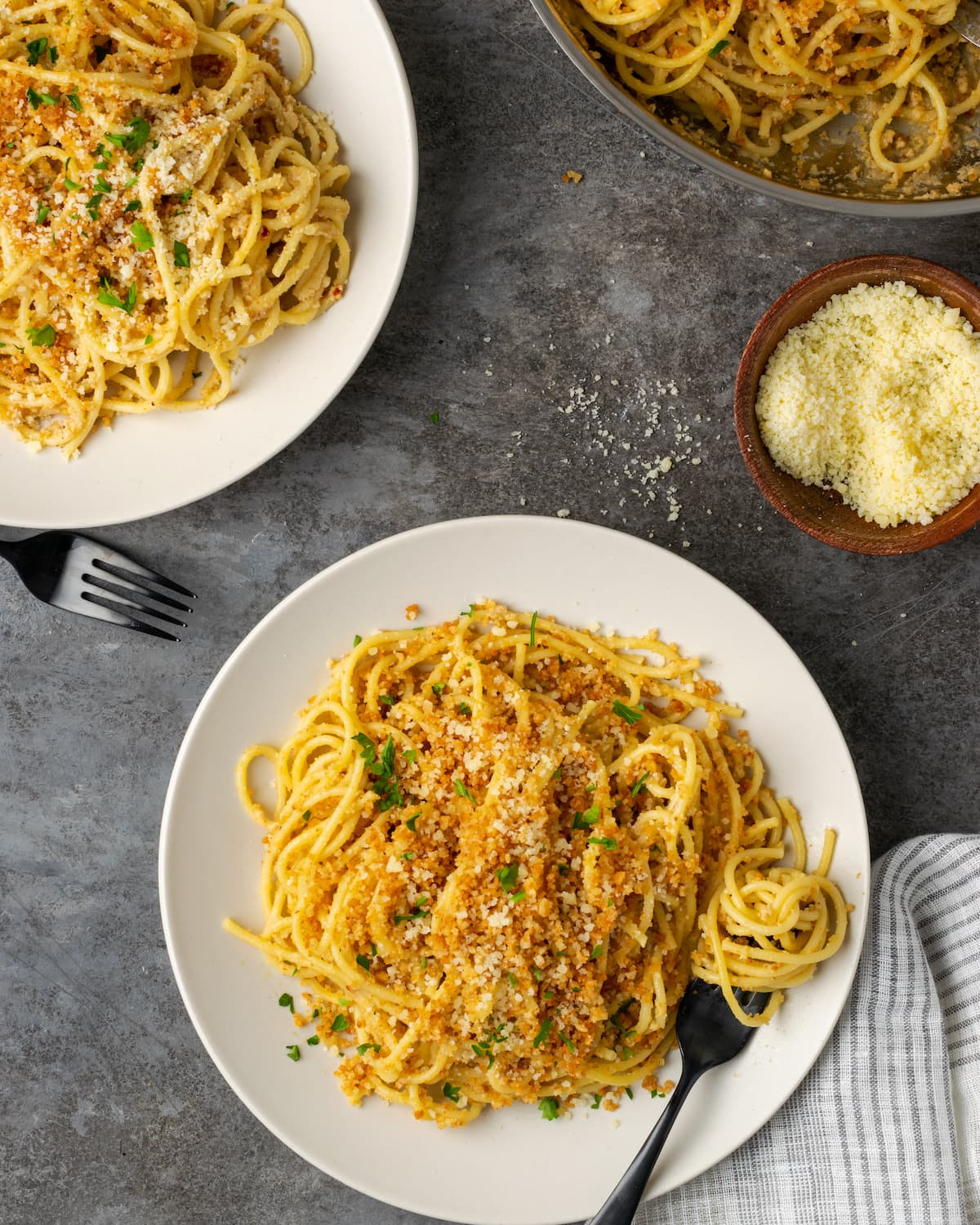 Vista aérea de dos platos de pasta César cubiertos con pan rallado con ajo, junto a una sartén y un tazón de queso parmesano rallado.
