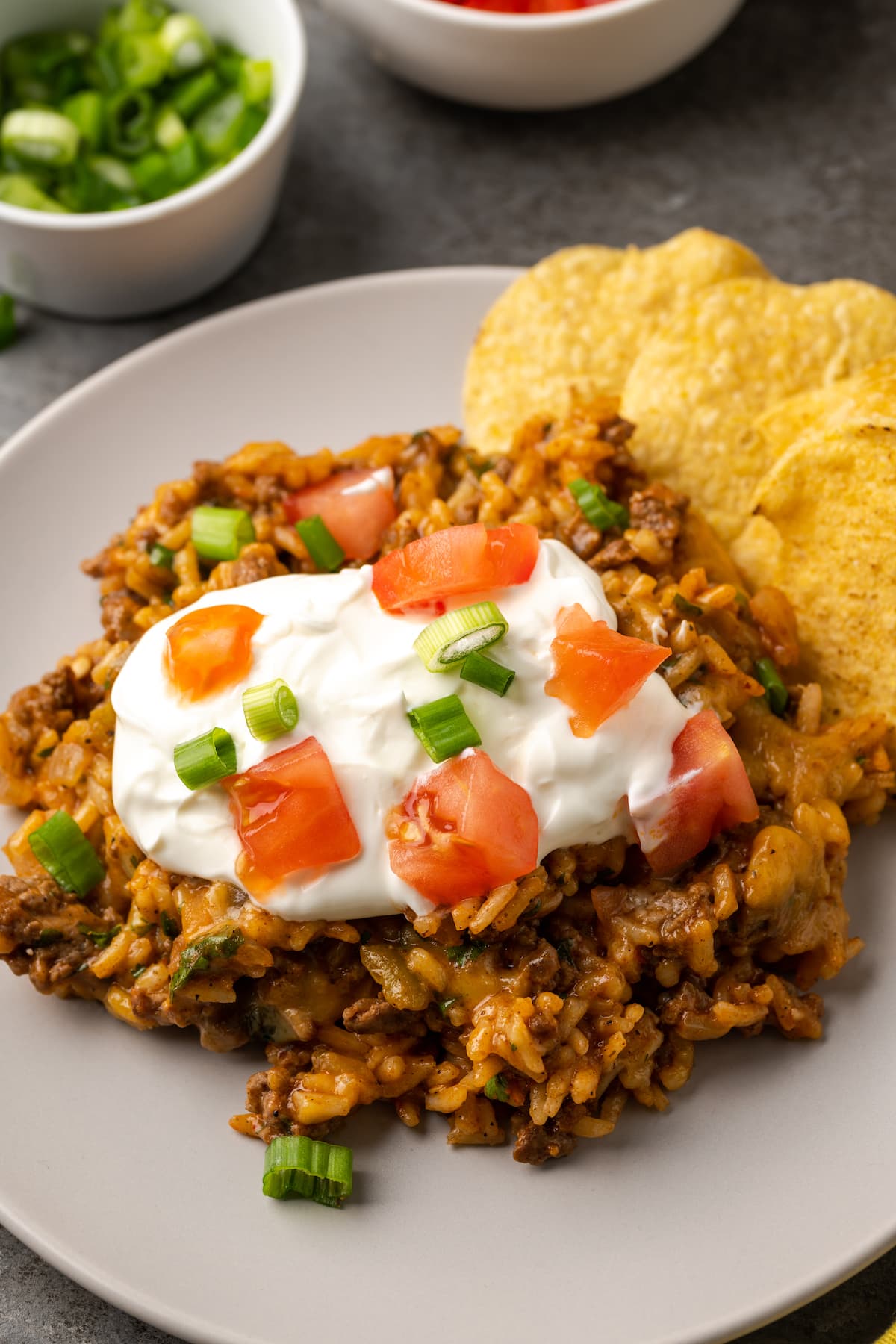 Una porción de taco de arroz con queso en un plato cubierto con crema agria, tomates cortados en cubitos y cebollas verdes junto a chips de maíz.