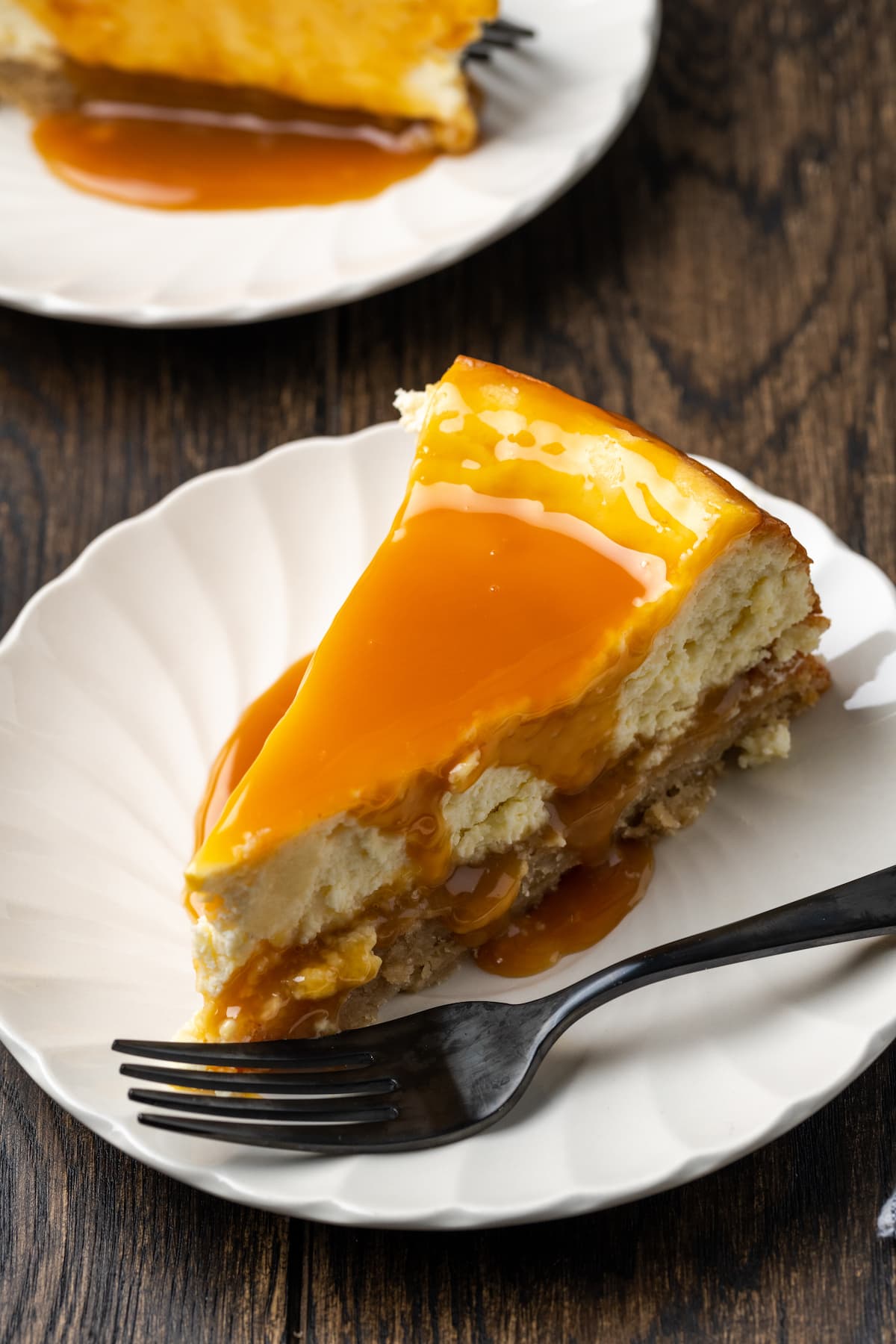 Banana Blondie Cheesecake es un postre fácil con cheesecake de vainilla al horno empapado en caramelo salado, en capas sobre una gruesa banana blondie. Un trozo de tarta de queso de banana blondie en un plato blanco con un tenedor, cubierto con caramelo salado.