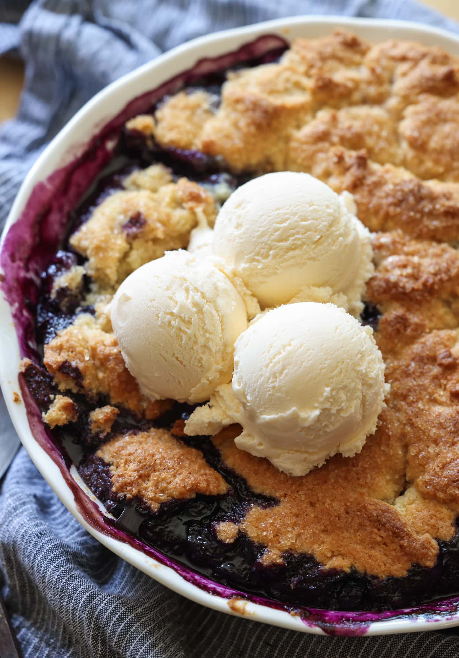 ¡Esta receta fácil de zapatero de arándanos es un postre clásico de verano que debes probar! Los arándanos dulces combinados con una cubierta de galleta mantecosa se hornean hasta que estén burbujeantes y dorados. Zapatero de arándanos en un molde para pastel con 3 bolas de helado de vainilla encima
