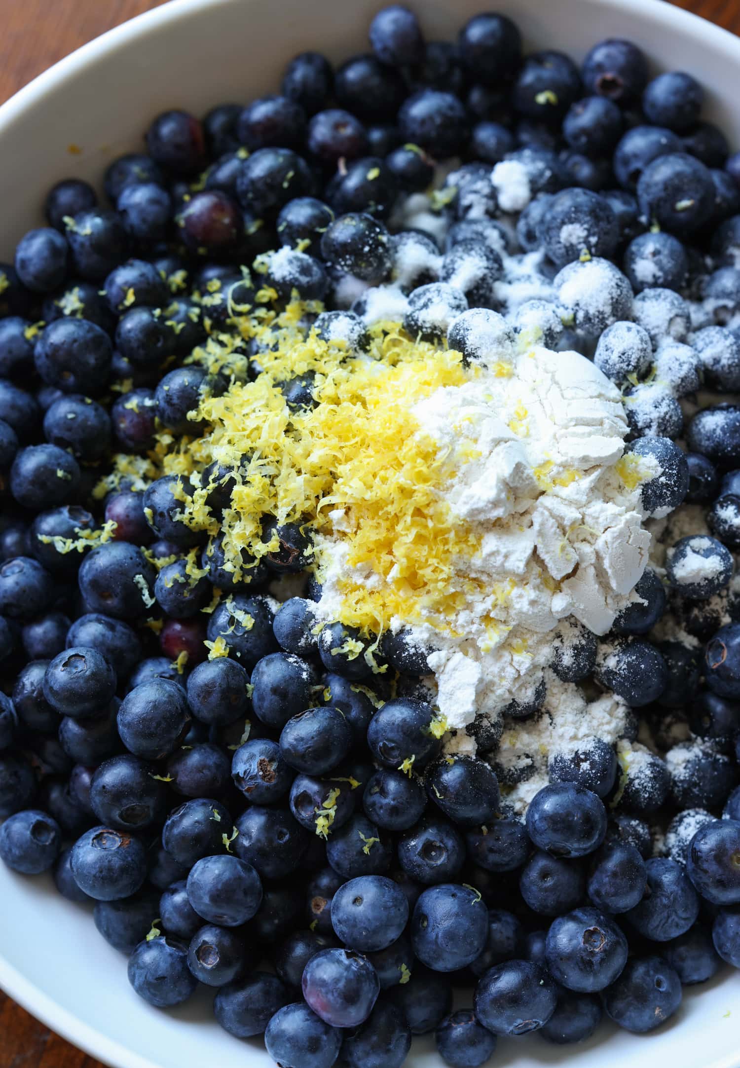 ¡Esta receta fácil de zapatero de arándanos es un postre clásico de verano que debes probar! Los arándanos dulces combinados con una cubierta de galleta mantecosa se hornean hasta que estén burbujeantes y dorados. Arándanos, harina y ralladura de limón en un tazón blanco