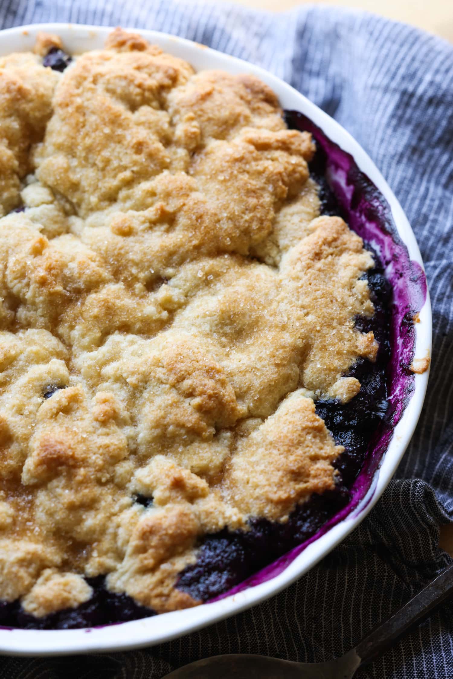 ¡Esta receta fácil de zapatero de arándanos es un postre clásico de verano que debes probar! Los arándanos dulces combinados con una cubierta de galleta mantecosa se hornean hasta que estén burbujeantes y dorados. un zapatero de arándanos en un plato de pastel blanco recién salido del horno