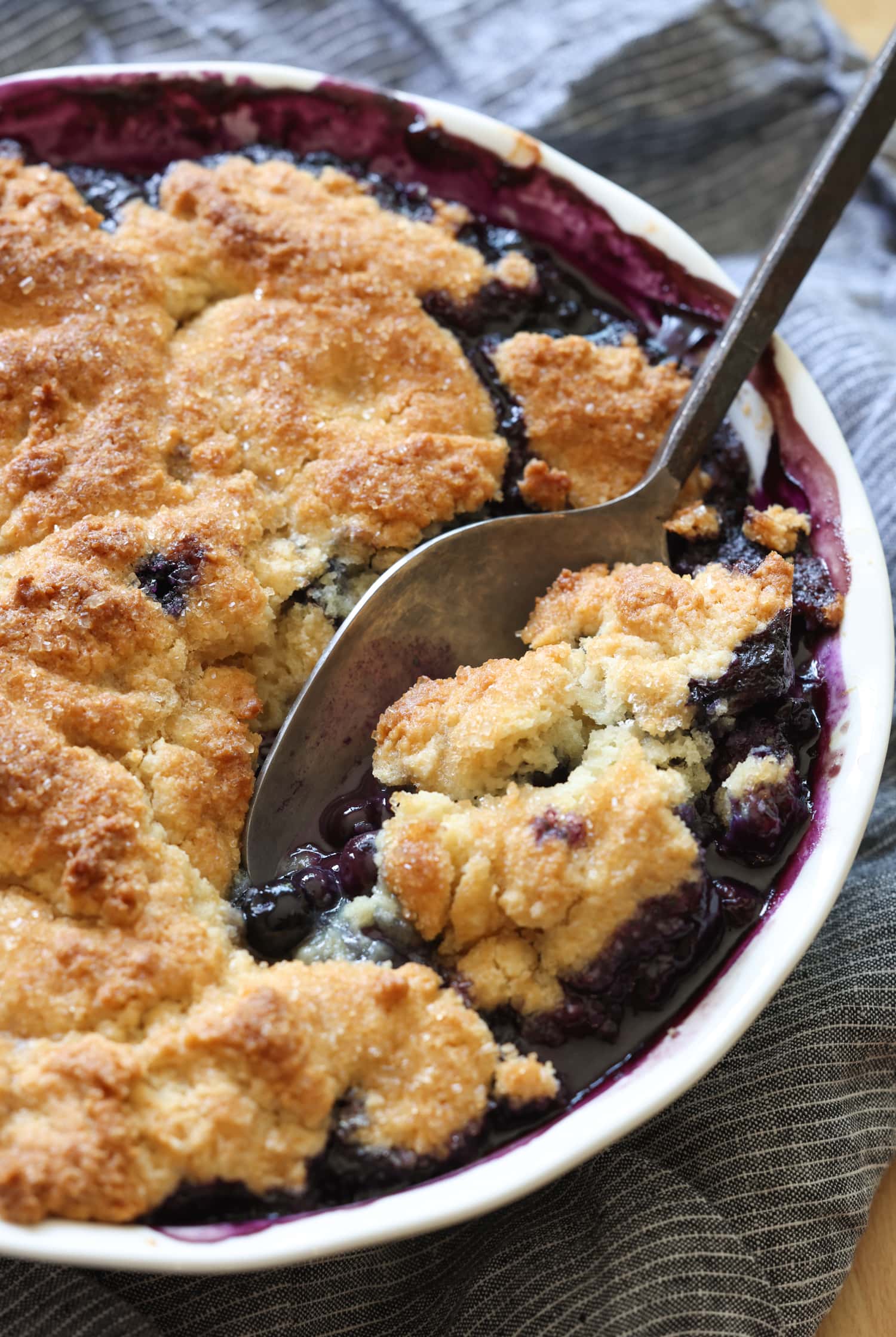 ¡Esta receta fácil de zapatero de arándanos es un postre clásico de verano que debes probar! Los arándanos dulces combinados con una cubierta de galleta mantecosa se hornean hasta que estén burbujeantes y dorados. Zapatero de arándanos en una fuente para horno con una cuchara sacando una porción