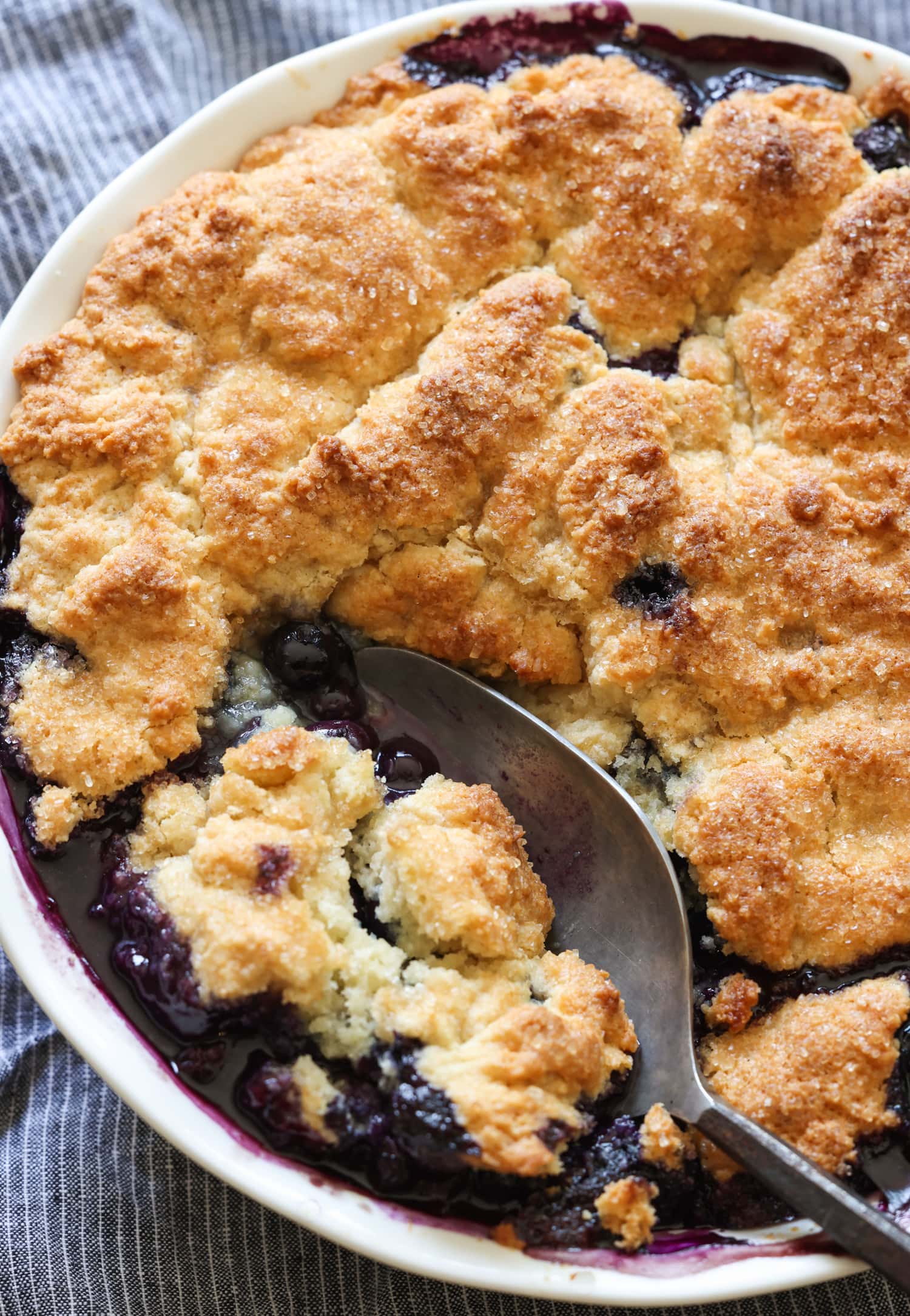 ¡Esta receta fácil de zapatero de arándanos es un postre clásico de verano que debes probar! Los arándanos dulces combinados con una cubierta de galleta mantecosa se hornean hasta que estén burbujeantes y dorados. Zapatero en una fuente para horno blanca con una cuchara que lo sirve.