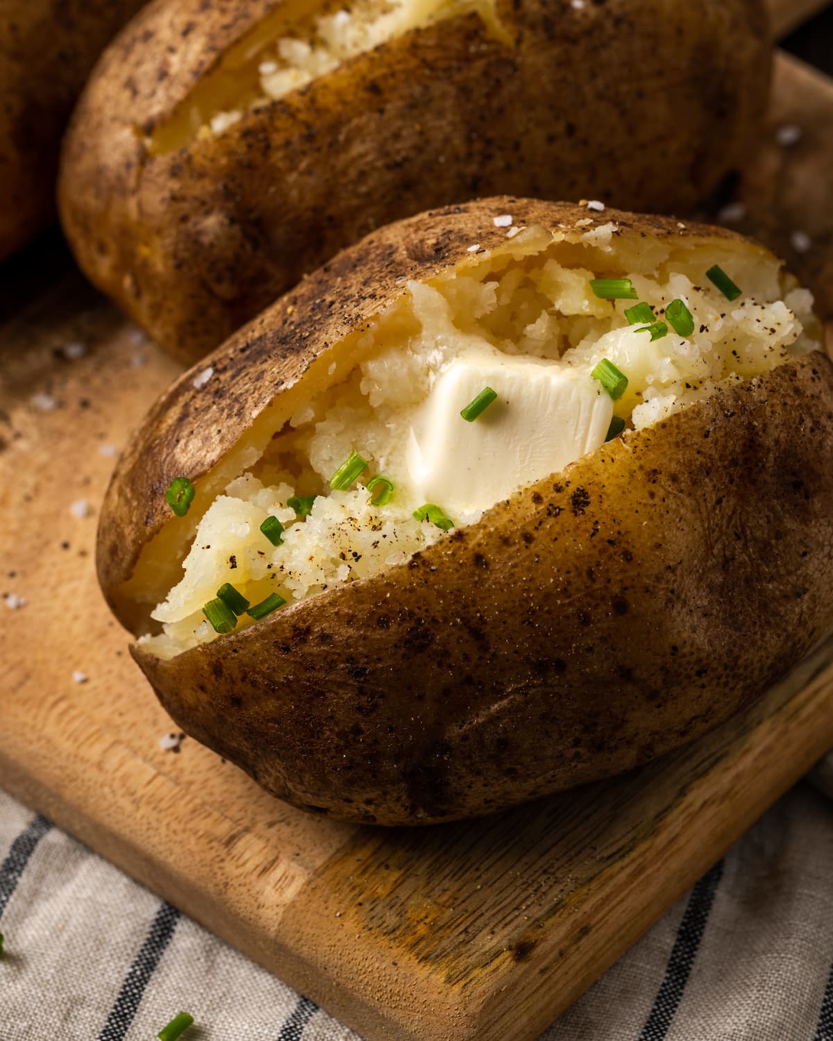 ¡Esta receta de papas al horno Crock Pot es un método más fácil para cocinar su guarnición de vegetales favorita sin encender el horno! Primer plano de una patata al horno crock pot cubierta con mantequilla y cebollino en una tabla de madera.