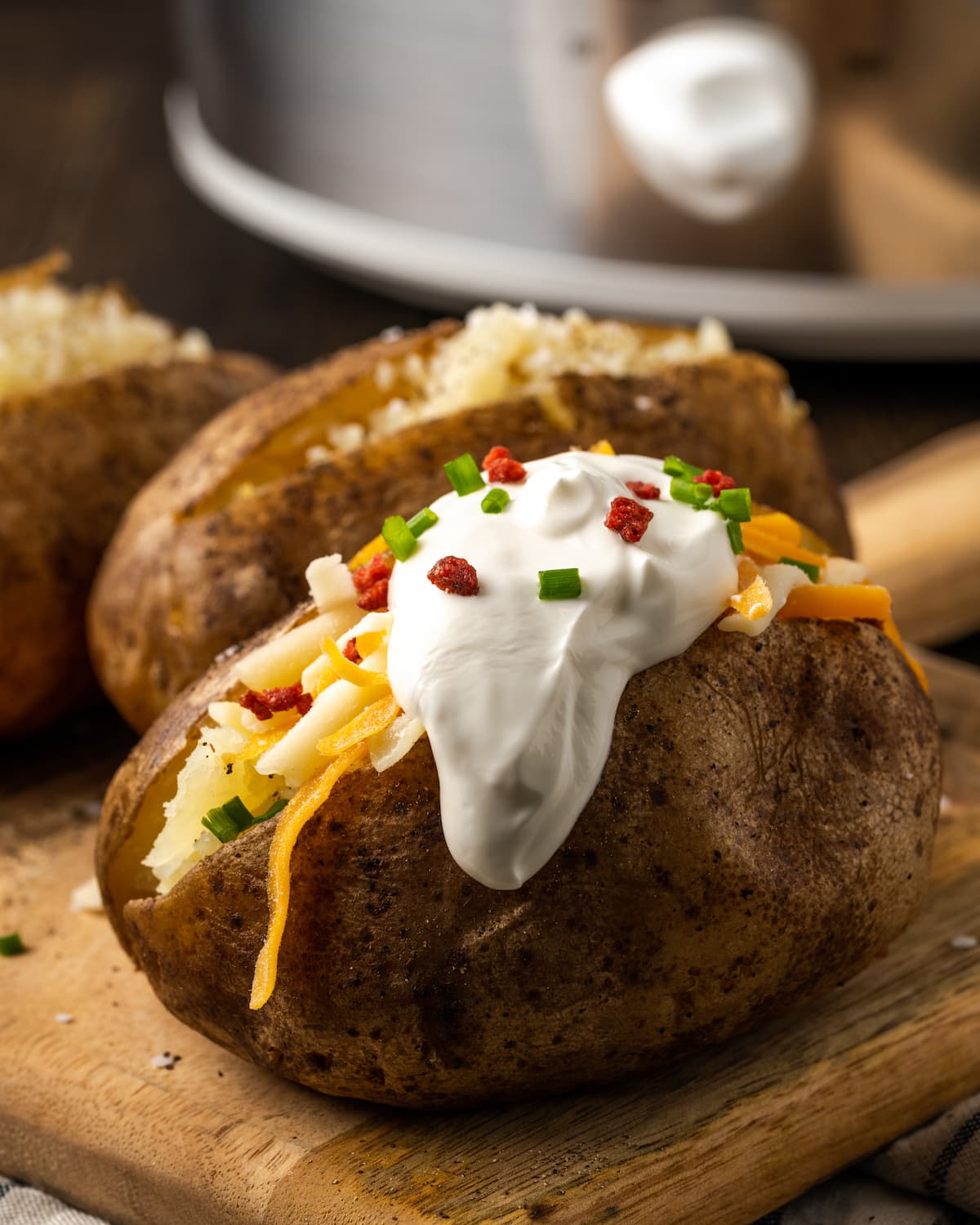 ¡Esta receta de papas al horno Crock Pot es un método más fácil para cocinar su guarnición de vegetales favorita sin encender el horno! Una papa horneada en olla de barro cubierta con crema agria, trocitos de tocino y cebollín sobre una tabla de madera.