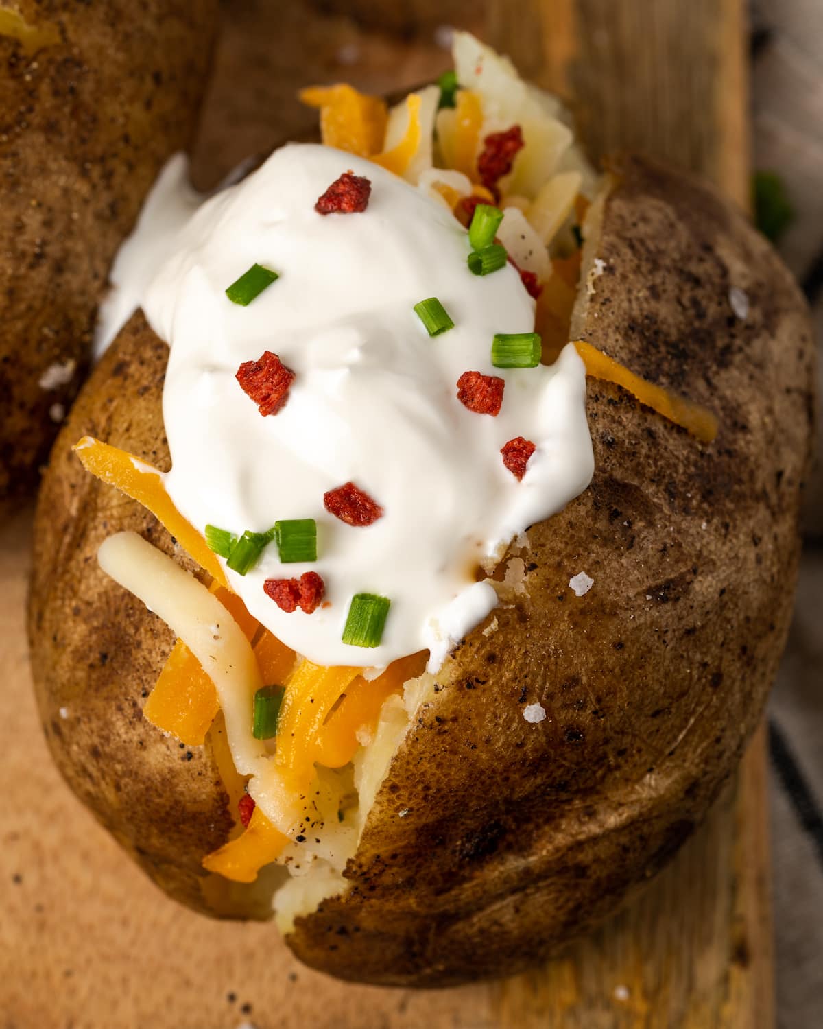 ¡Esta receta de papas al horno Crock Pot es un método más fácil para cocinar su guarnición de vegetales favorita sin encender el horno! Primer plano de una patata al horno de olla de barro cubierta con crema agria, trocitos de tocino y cebollino en una tabla de madera.
