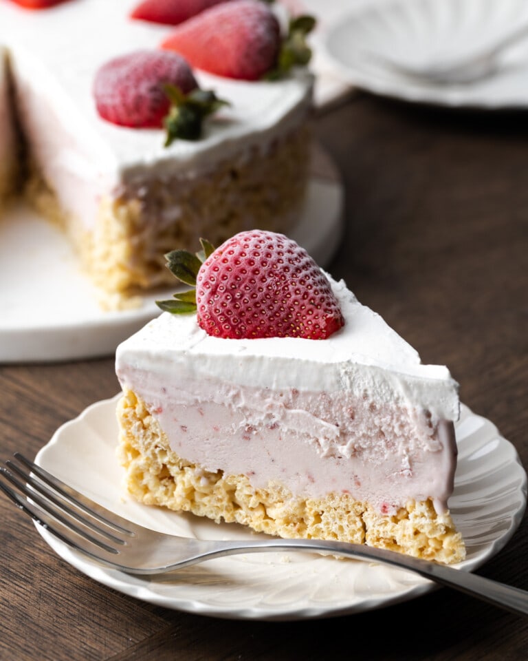 Un trozo de tarta de helado Krispie Treat en un plato blanco junto a un tenedor, adornado con una fresa fresca, con el resto de la tarta en el fondo.