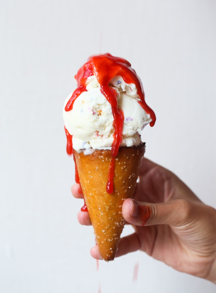 Una mano que sostiene un cono de helado de vainilla que gotea con jarabe de fresa.