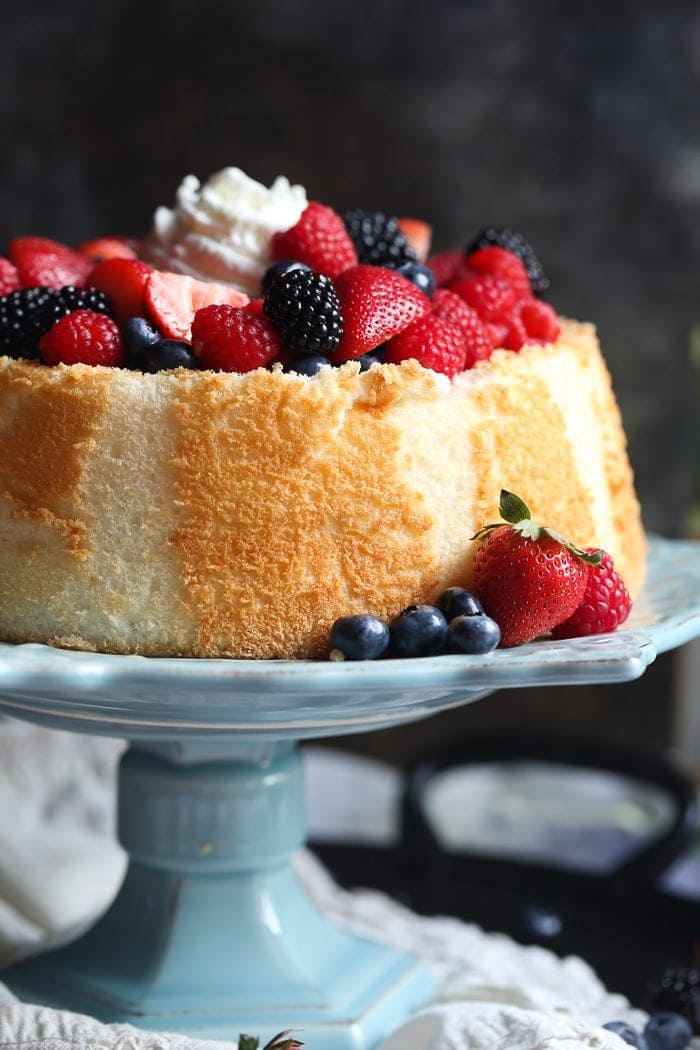 Angel Food Cake cubierto con bayas mixtas empapadas en vino sobre un puesto de pastel azul.