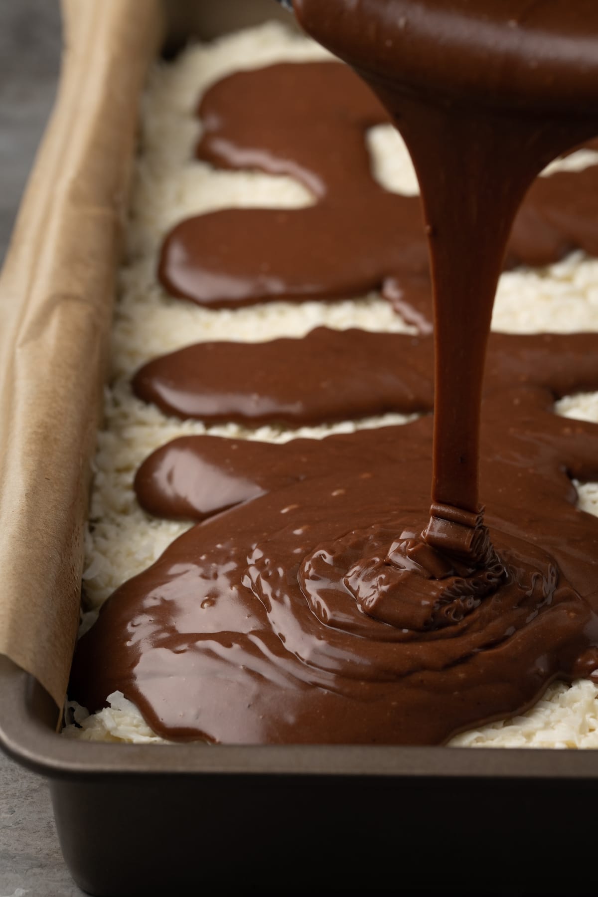 El glaseado de chocolate se vierte sobre la parte superior de las capas de brownie y coco en un molde para hornear forrado con pergamino.