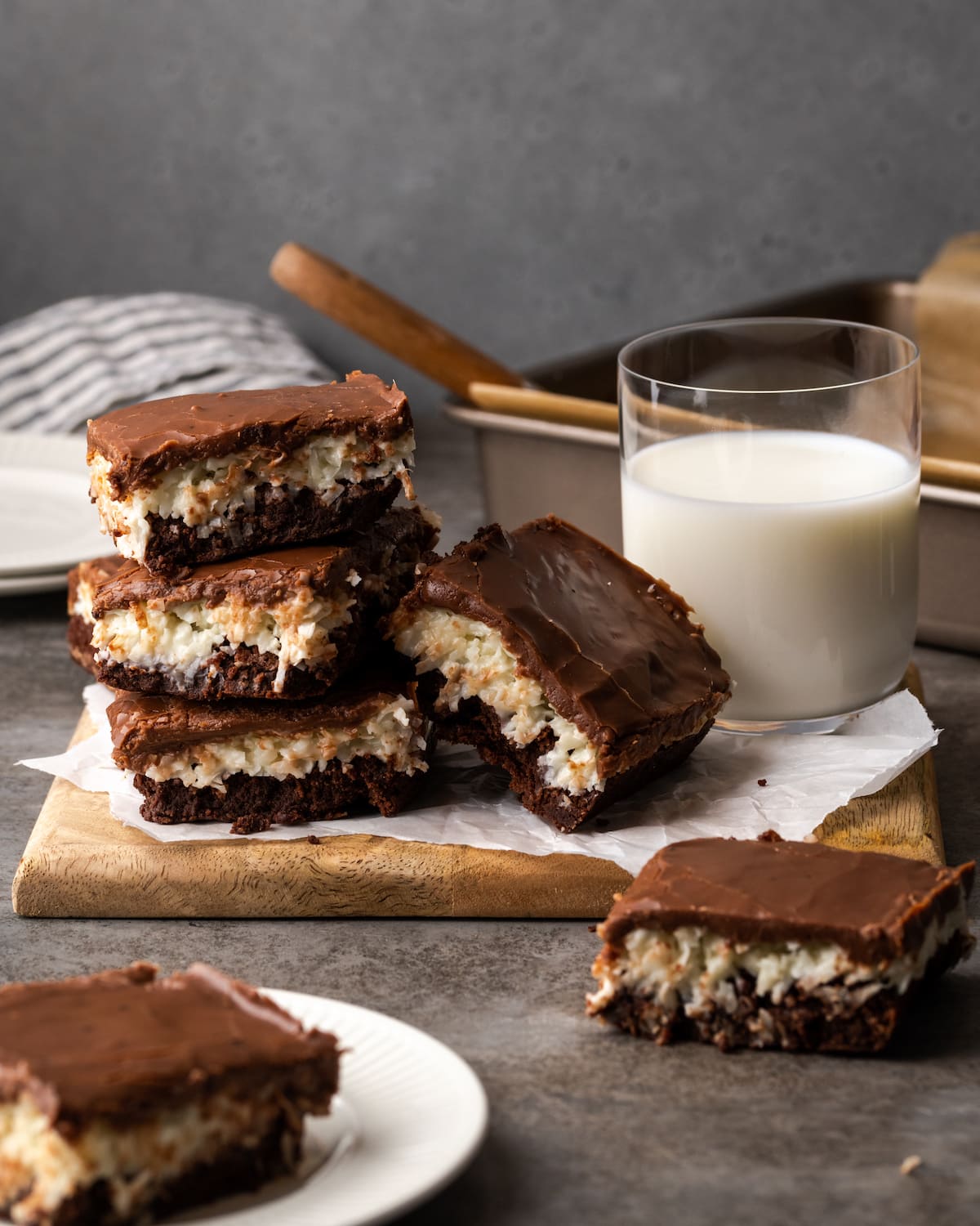 Una pila de brownies de coco en una fuente de madera junto a un vaso de leche, con una fuente de brownies al fondo.