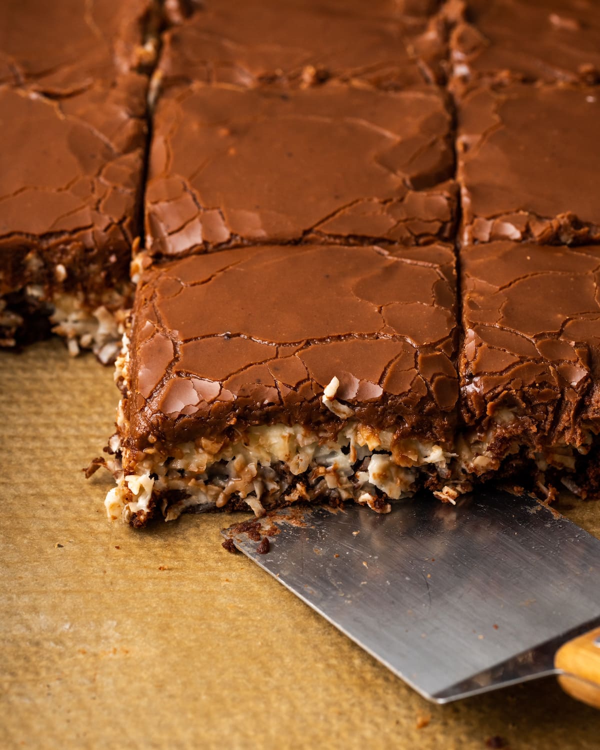 Una espátula desplazada se desliza debajo de los brownies de coco en una fuente forrada.