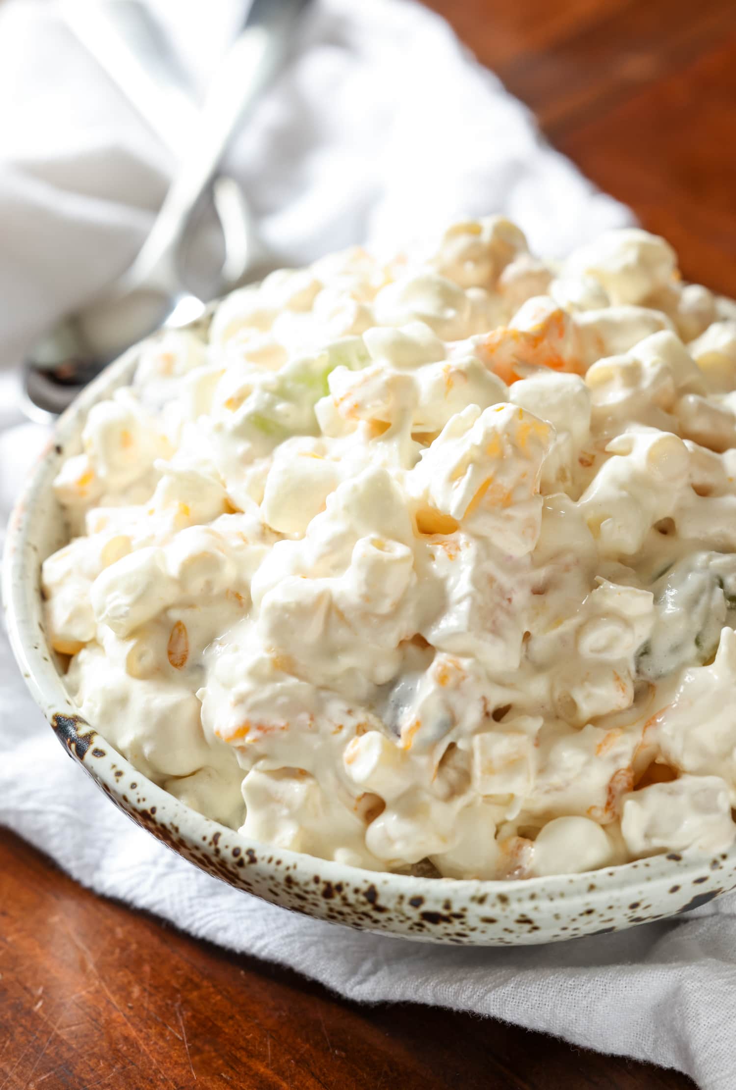 Frog Eye Salad es una ensalada dulce de verano con fruta, natillas, cobertura batida y pasta. ¡Es una delicia de textura y sabor, y es la receta de guarnición perfecta para su barbacoa de verano! Ensalada de ojo de rana en un recipiente para servir sobre un paño de cocina blanco