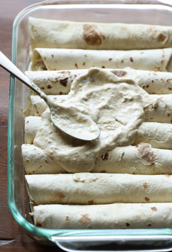 Cuchara de salsa verde con salsa de crema agria encima de enchiladas enrolladas en una sartén de 9x13