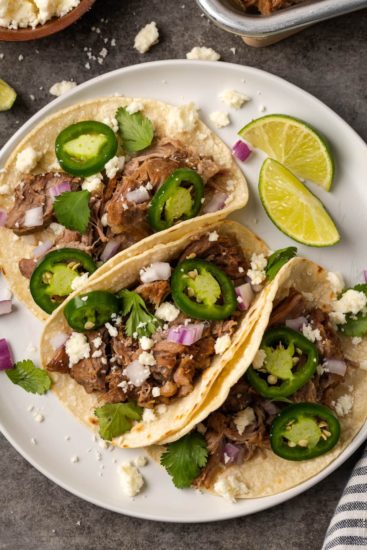 ¡Esta receta de Instant Pot Carnitas necesita solo 5 minutos de preparación! Haga carnitas increíblemente tiernas, jugosas y crujientes para tacos de cerdo y más. Vista aérea de tres tacos de cerdo cubiertos con jalapeños y queso en un plato blanco junto a rodajas de lima.