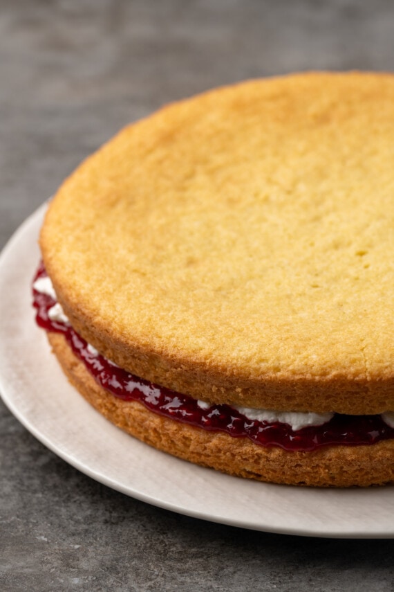 Esta receta de bizcocho Victoria es suave, esponjosa y está hecha con ingredientes simples. ¡Perfecto para combinar con mermelada y crema batida casera! Capas de bizcocho ensambladas rellenas de mermelada de frambuesa y crema batida en un plato.