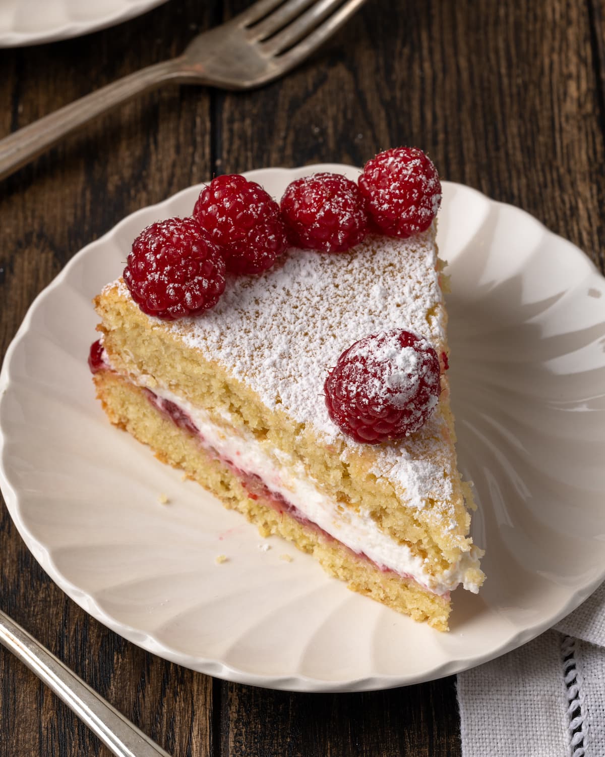 Esta receta de bizcocho Victoria es suave, esponjosa y está hecha con ingredientes simples. ¡Perfecto para combinar con mermelada y crema batida casera! Un trozo de bizcocho Victoria en un plato blanco, cubierto con frambuesas frescas.