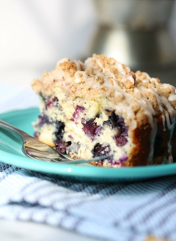 Una rebanada de pastel de muffin de arándanos junto a un tenedor en un plato turquesa.