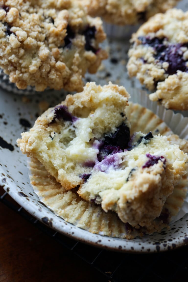 Un muffin de arándanos se abrió para revelar el relleno de queso crema, junto a más muffins en un plato.