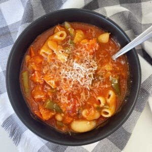 Vista aérea de la sopa minestrone en un tazón negro cubierto con queso parmesano rallado. El tazón está sobre un paño de cocina a cuadros en blanco y negro.