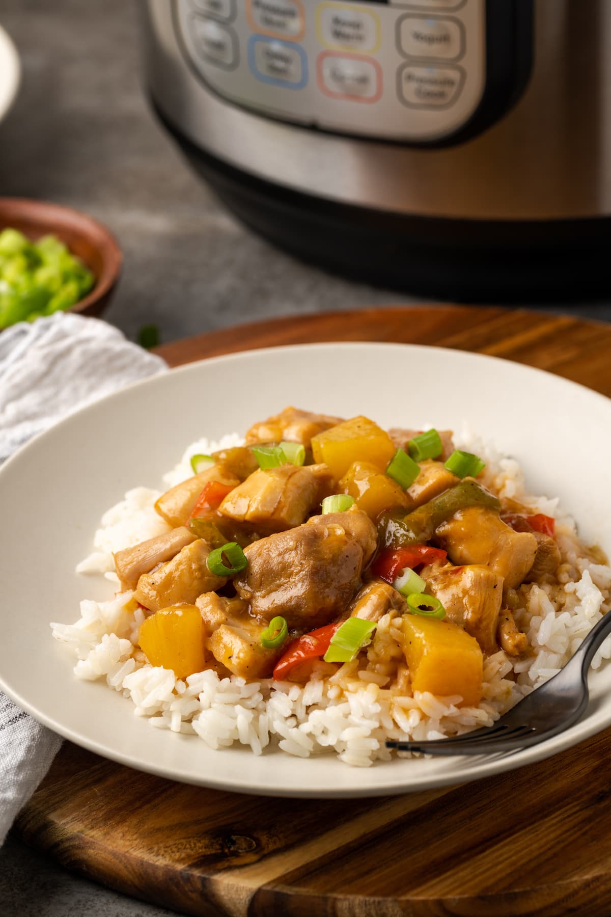 Instant Pot Hawaiian Chicken es una cena fácil de una olla cocinada con piña jugosa, pimientos y cebollas en una salsa dulce y picante. Pollo hawaiano servido sobre arroz en un plato blanco junto a un tenedor, con la olla instantánea al fondo.