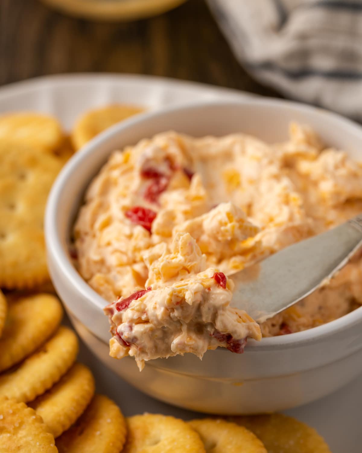 Esta receta cremosa de queso pimiento es un clásico aperitivo sureño para mojar y untar elaborado con queso cheddar y pimientos pimientos dulces. Primer plano de un plato de queso pimiento servido con un cuchillo de queso en un plato de galletas saladas.