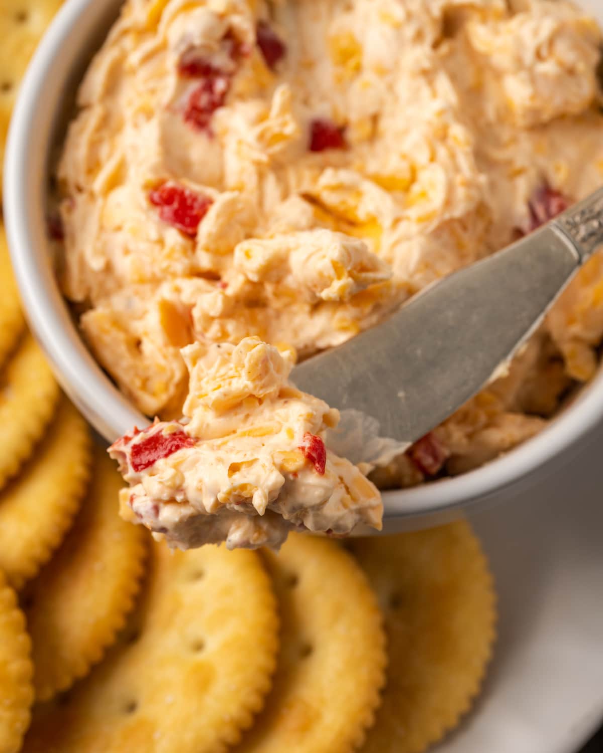 Esta receta cremosa de queso pimiento es un clásico aperitivo sureño para mojar y untar elaborado con queso cheddar y pimientos pimientos dulces. Primer plano de un plato de queso pimiento servido con un cuchillo de queso en un plato de galletas saladas.