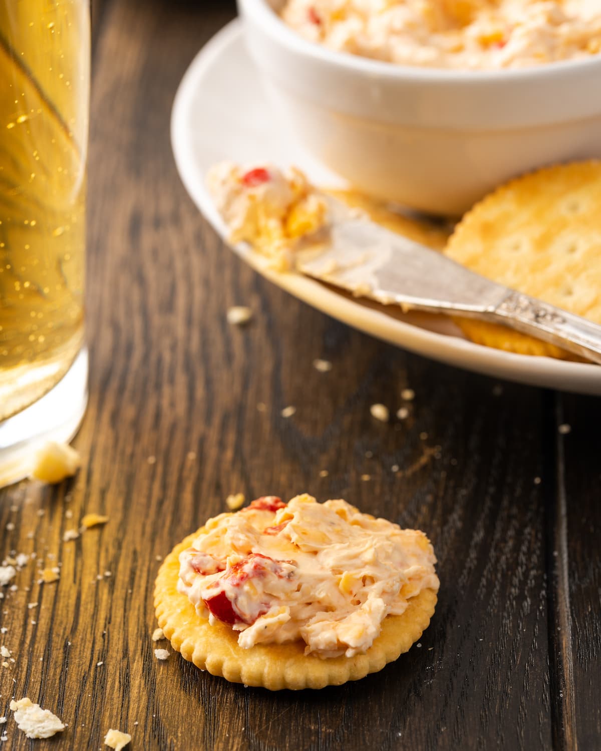 Esta receta cremosa de queso pimiento es un clásico aperitivo sureño para mojar y untar elaborado con queso cheddar y pimientos pimientos dulces. Una galleta cubierta con queso pimiento en una mesa con un plato de galletas y queso pimiento en el fondo.