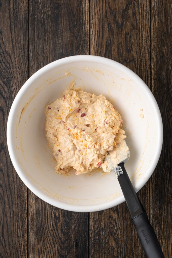 Esta receta cremosa de queso pimiento es un clásico aperitivo sureño para mojar y untar elaborado con queso cheddar y pimientos pimientos dulces. Queso pimiento revuelto en un tazón blanco con una espátula.