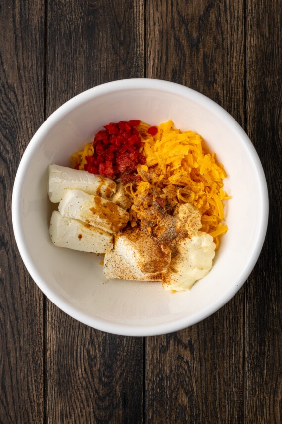 Esta receta cremosa de queso pimiento es un clásico aperitivo sureño para mojar y untar elaborado con queso cheddar y pimientos pimientos dulces. Ingredientes de queso pimiento combinados en un tazón blanco.