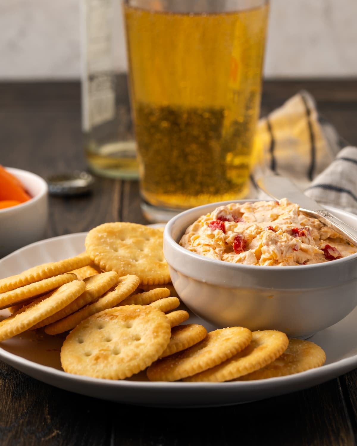 Esta receta cremosa de queso pimiento es un clásico aperitivo sureño para mojar y untar elaborado con queso cheddar y pimientos pimientos dulces. Un plato de queso pimiento servido en un plato de galletas saladas, con vasos de cerveza de fondo.
