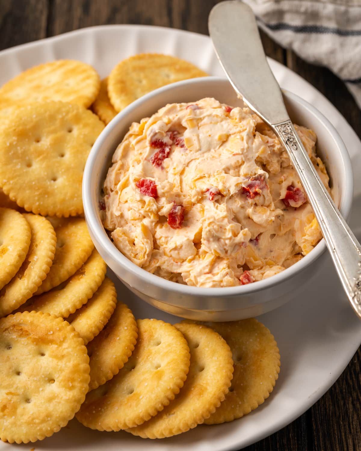 Esta receta cremosa de queso pimiento es un clásico aperitivo sureño para mojar y untar elaborado con queso cheddar y pimientos pimientos dulces. Un plato de queso pimiento servido con un cuchillo de queso en un plato de galletas saladas.