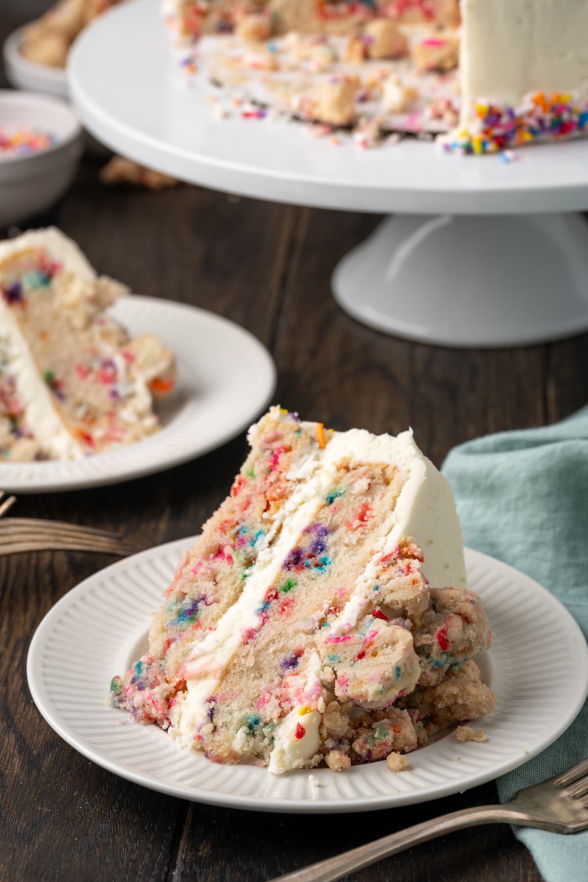 This is the best confetti cake recipe made from fluffy white cake loaded with rainbow sprinkles. Even better than a boxed Funfetti cake mix! A slice of frosted confetti cake on a white plate, with a second slice on a plate next to a cake stand in the background.