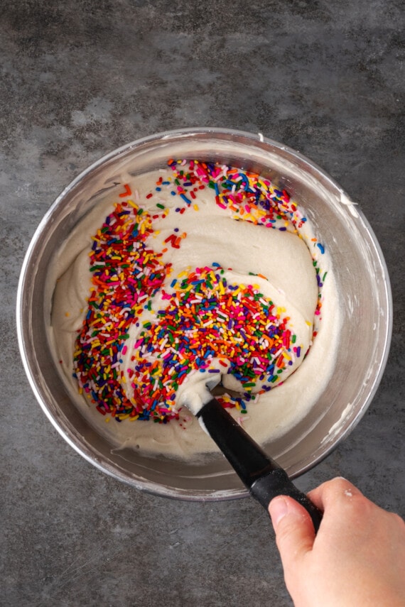 This is the best confetti cake recipe made from fluffy white cake loaded with rainbow sprinkles. Even better than a boxed Funfetti cake mix! A hand using a black spoon to stir rainbow sprinkles into cake batter in a metal bowl.