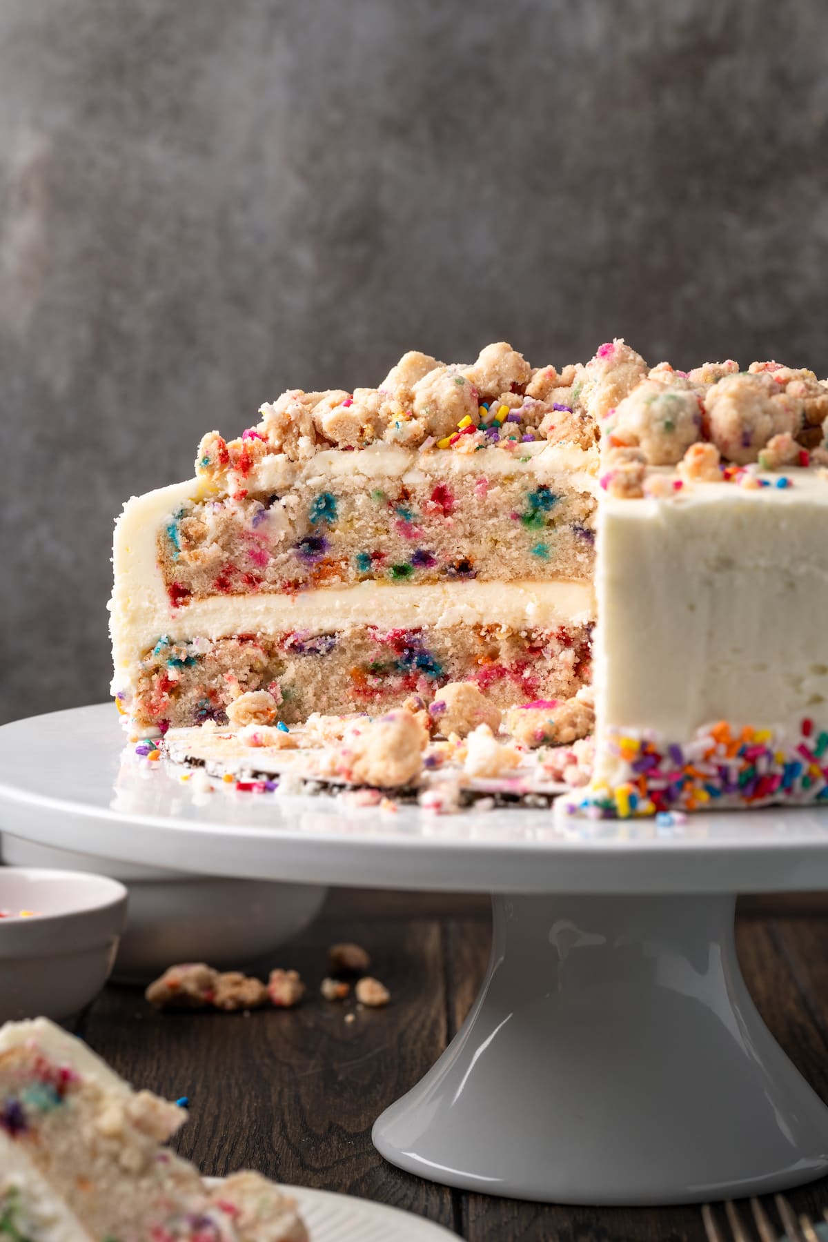 This is the best confetti cake recipe made from fluffy white cake loaded with rainbow sprinkles. Even better than a boxed Funfetti cake mix! Frosted confetti cake on a cake stand topped with funfetti streusel and sprinkles, with a large slice missing.