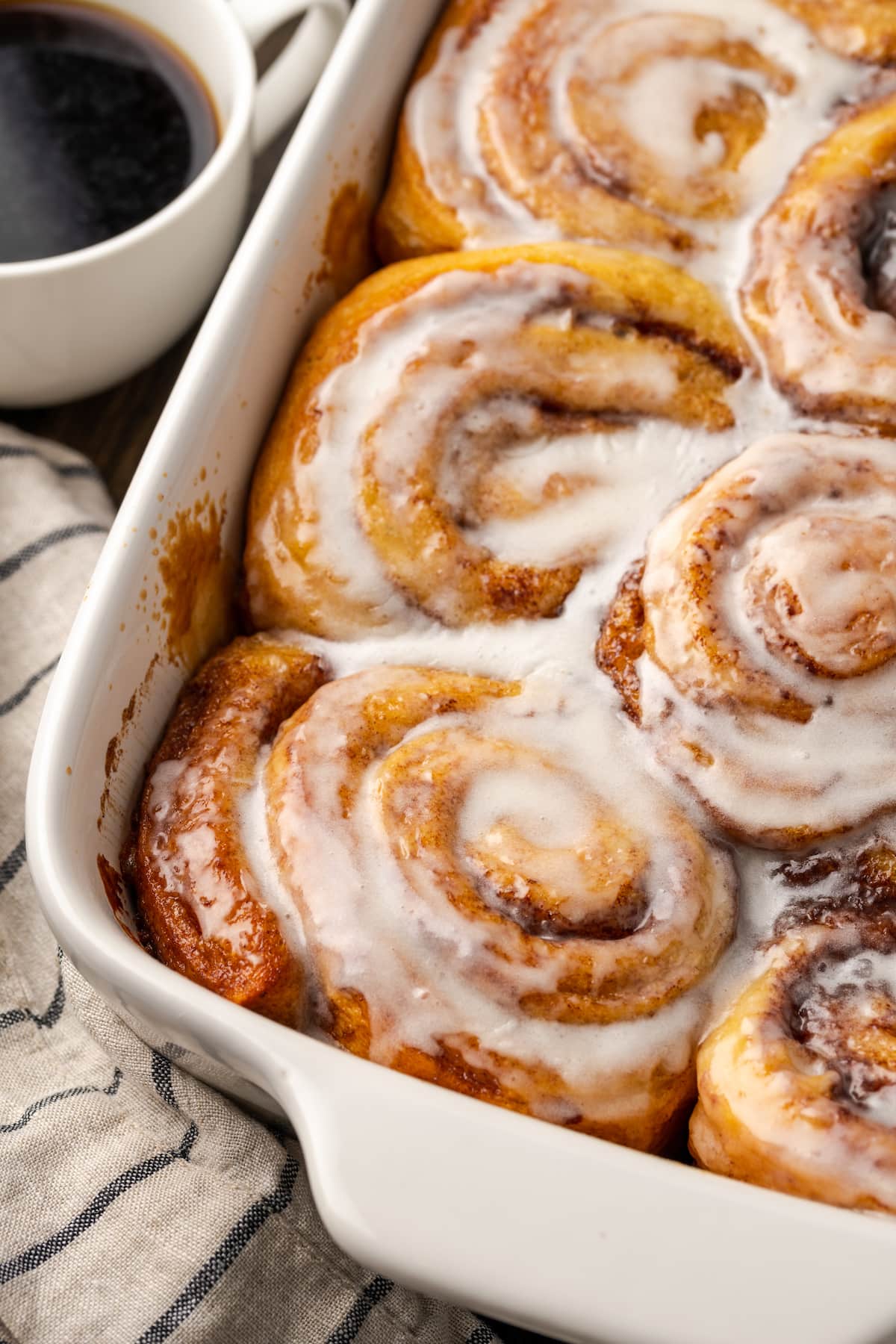 Make the viral TikTok cinnamon rolls with heavy cream! These easy cinnamon rolls taste homemade, perfect as a sweet breakfast or easy dessert. Overhead view of baked TikTok cinnamon rolls covered with icing in a ceramic baking dish, next to a cup of coffee.