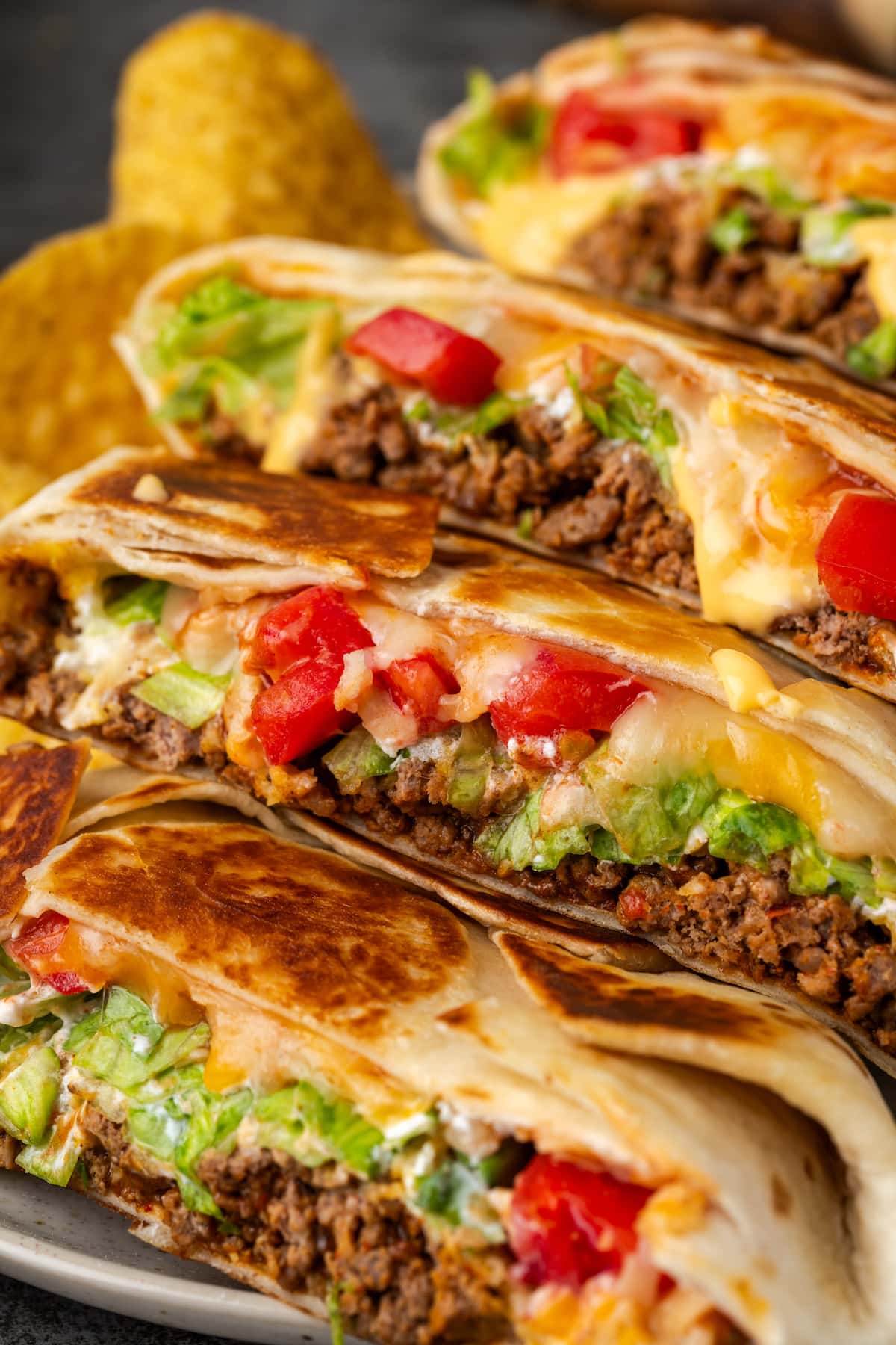 Make satisfying crunch wraps loaded with taco meat, melty cheese, tostada, sour cream, and more wrapped up inside a toasty flour tortilla! Close up of crunch wraps cut in half and arranged in a row on a plate next to corn chips.