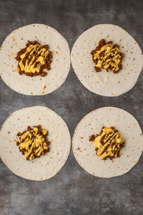 Make satisfying crunch wraps loaded with taco meat, melty cheese, tostada, sour cream, and more wrapped up inside a toasty flour tortilla! Overhead view of four partially assembled crunch wraps topped with meat and nacho cheese.