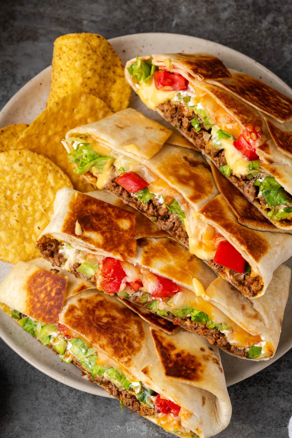 Make satisfying crunch wraps loaded with taco meat, melty cheese, tostada, sour cream, and more wrapped up inside a toasty flour tortilla! Crunch wraps cut in half and arranged in a row on a plate next to corn chips.