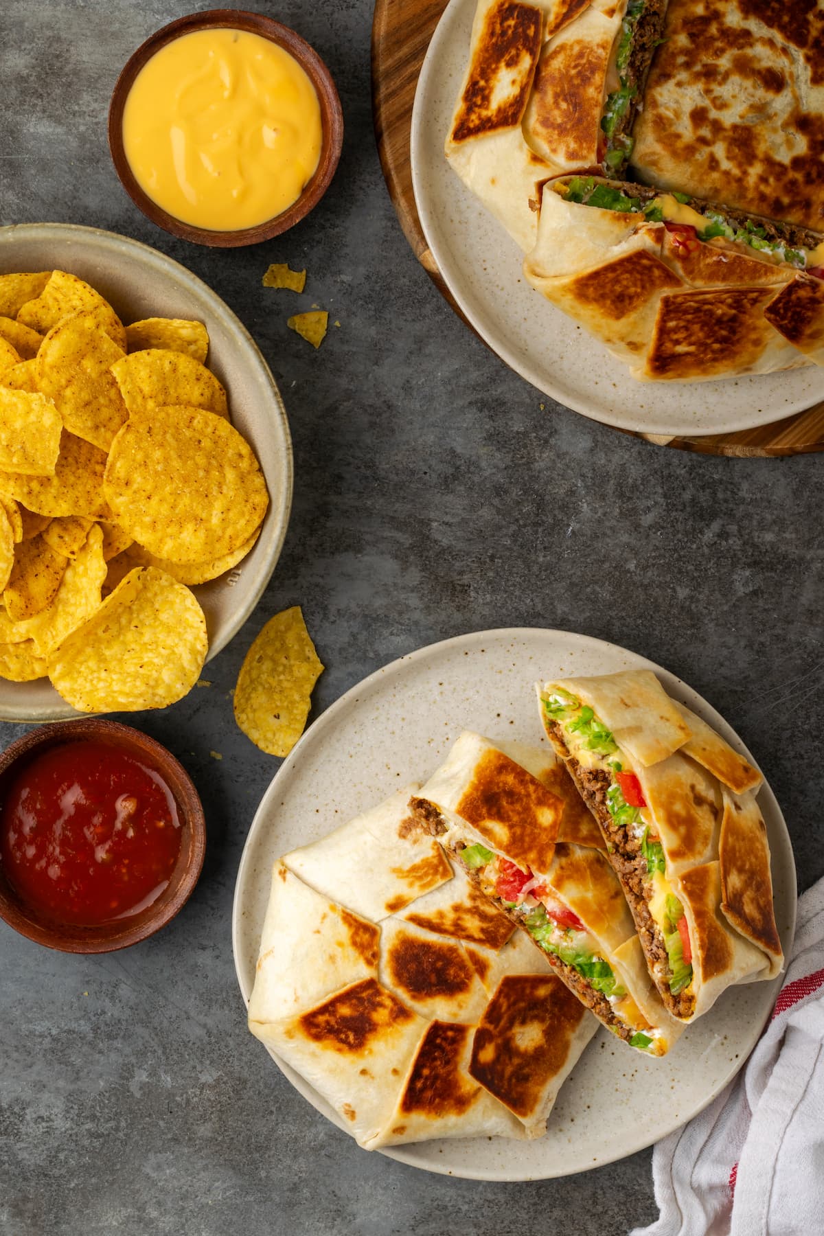 Make satisfying crunch wraps loaded with taco meat, melty cheese, tostada, sour cream, and more wrapped up inside a toasty flour tortilla! Overhead view of crunch wraps served on plates next to a bowl of corn chips and salsa for dipping.