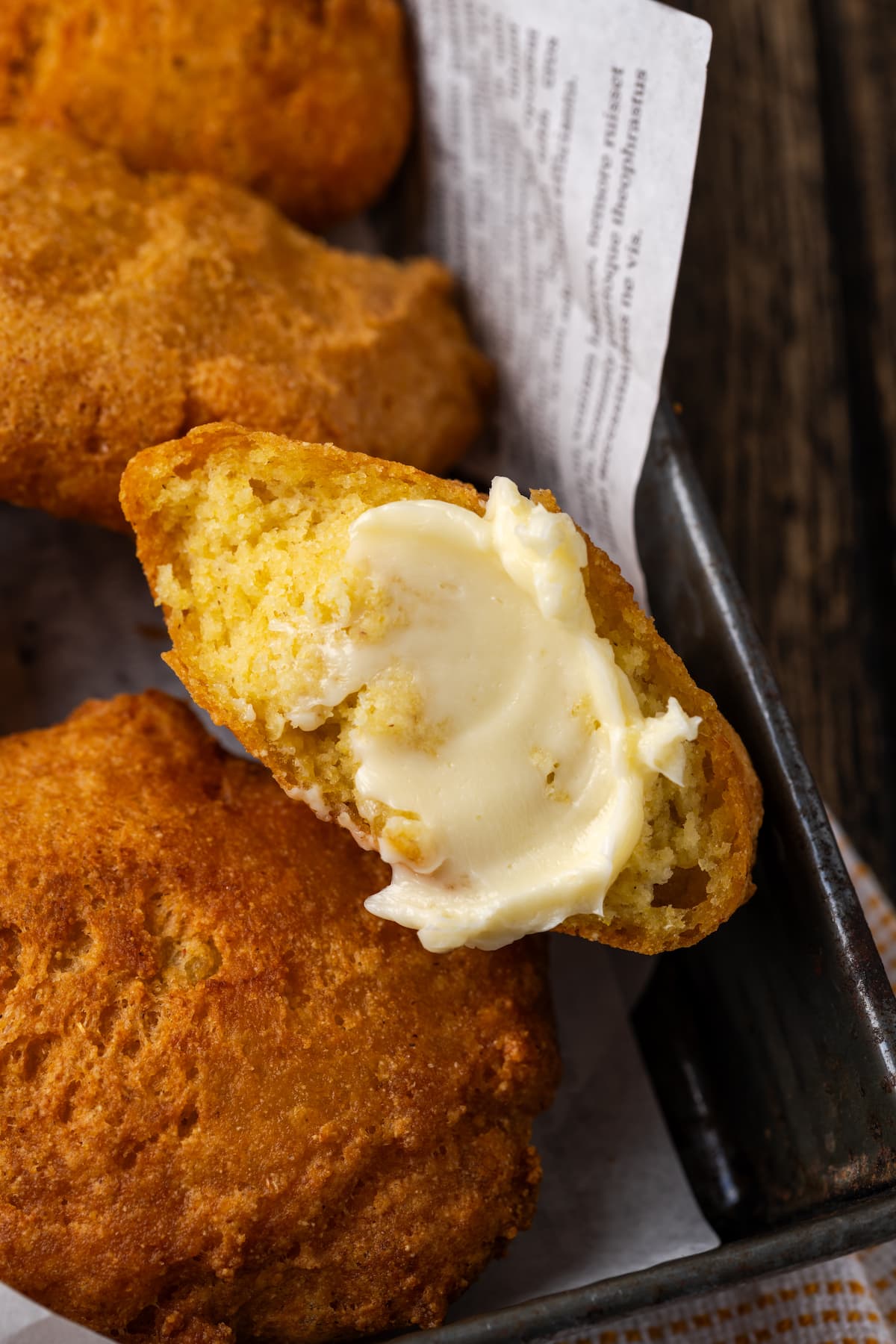 Crispy hot water cornbread is totally crave-worthy! Fried cornbread is an easy side dish to pair with your favorite Southern meals. Close up of hot water cornbread in a basket, with one fritter broken in half and spread with butter.