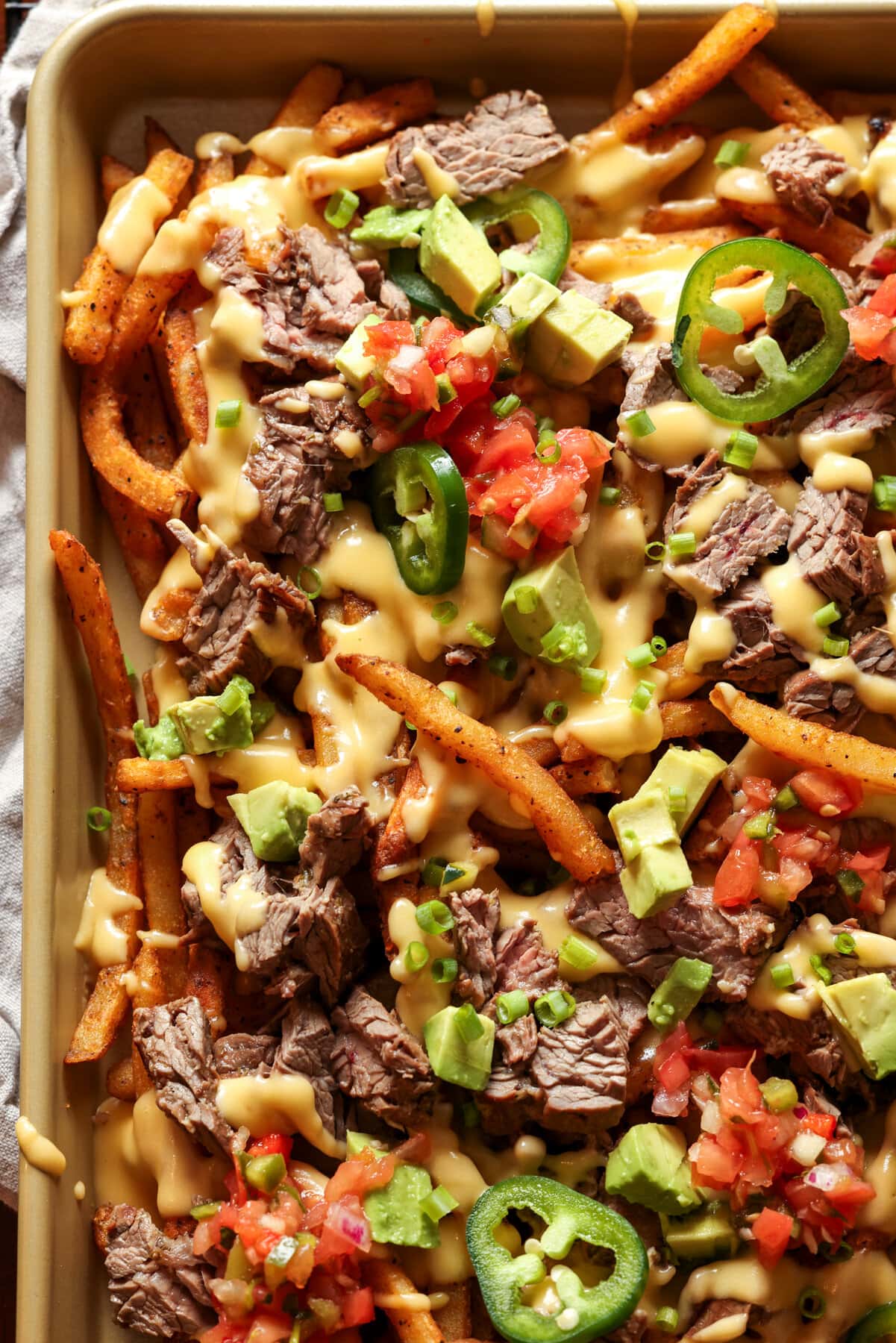 Carne Asada Fries Are a Tex Mex spin on loaded french fries. Marinated, grilled carne asada is sliced and served on top of seasoned fries with a cheesy sauce and all the toppings of your choice! Carne Asada Fries topped with melted cheese and all the fixings on a baking sheet