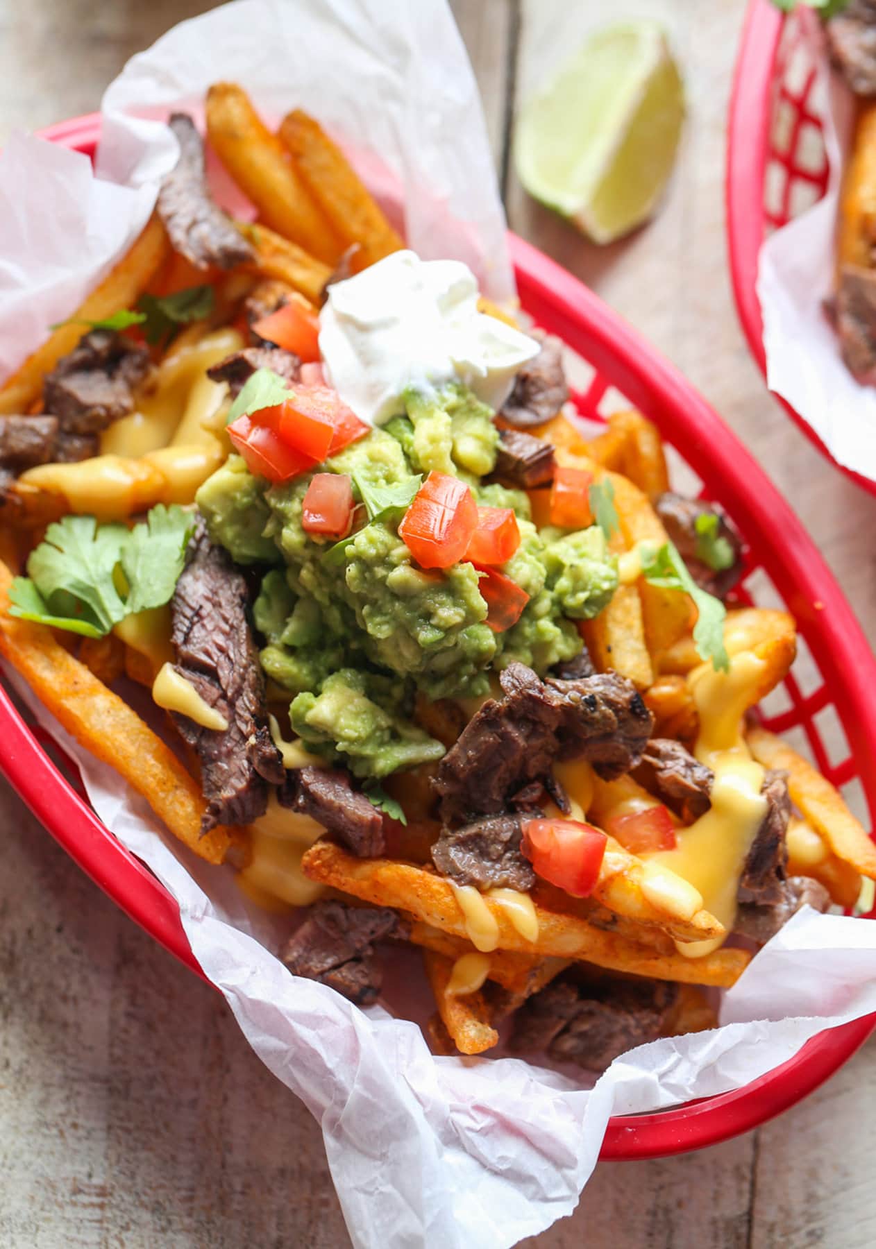 Carne Asada Fries Are a Tex Mex spin on loaded french fries. Marinated, grilled carne asada is sliced and served on top of seasoned fries with a cheesy sauce and all the toppings of your choice! Carne Asada French Fries topped with guacamole, cheese sauce, and sour cream in a basket