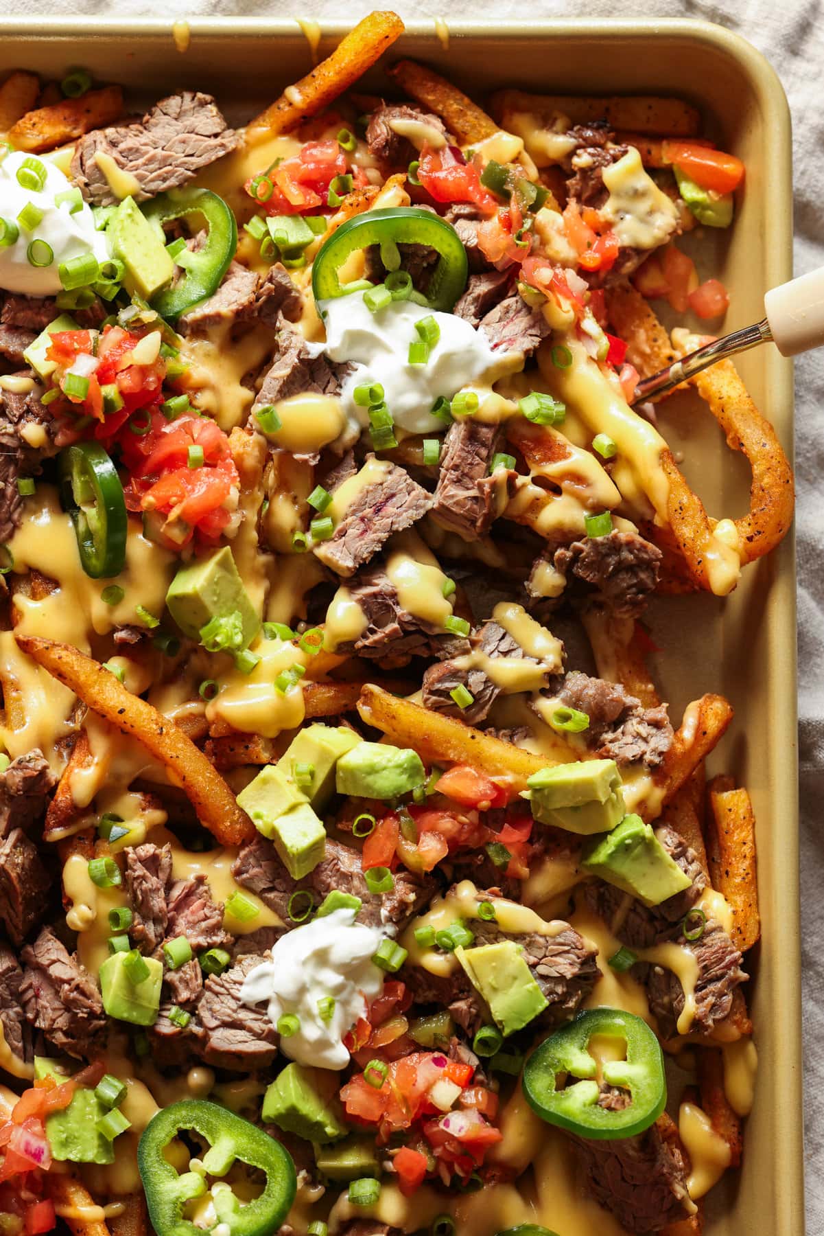 Carne Asada Fries Are a Tex Mex spin on loaded french fries. Marinated, grilled carne asada is sliced and served on top of seasoned fries with a cheesy sauce and all the toppings of your choice! Carne Asada Fries on a large baking sheet topped with sour cream and pico de gallo.