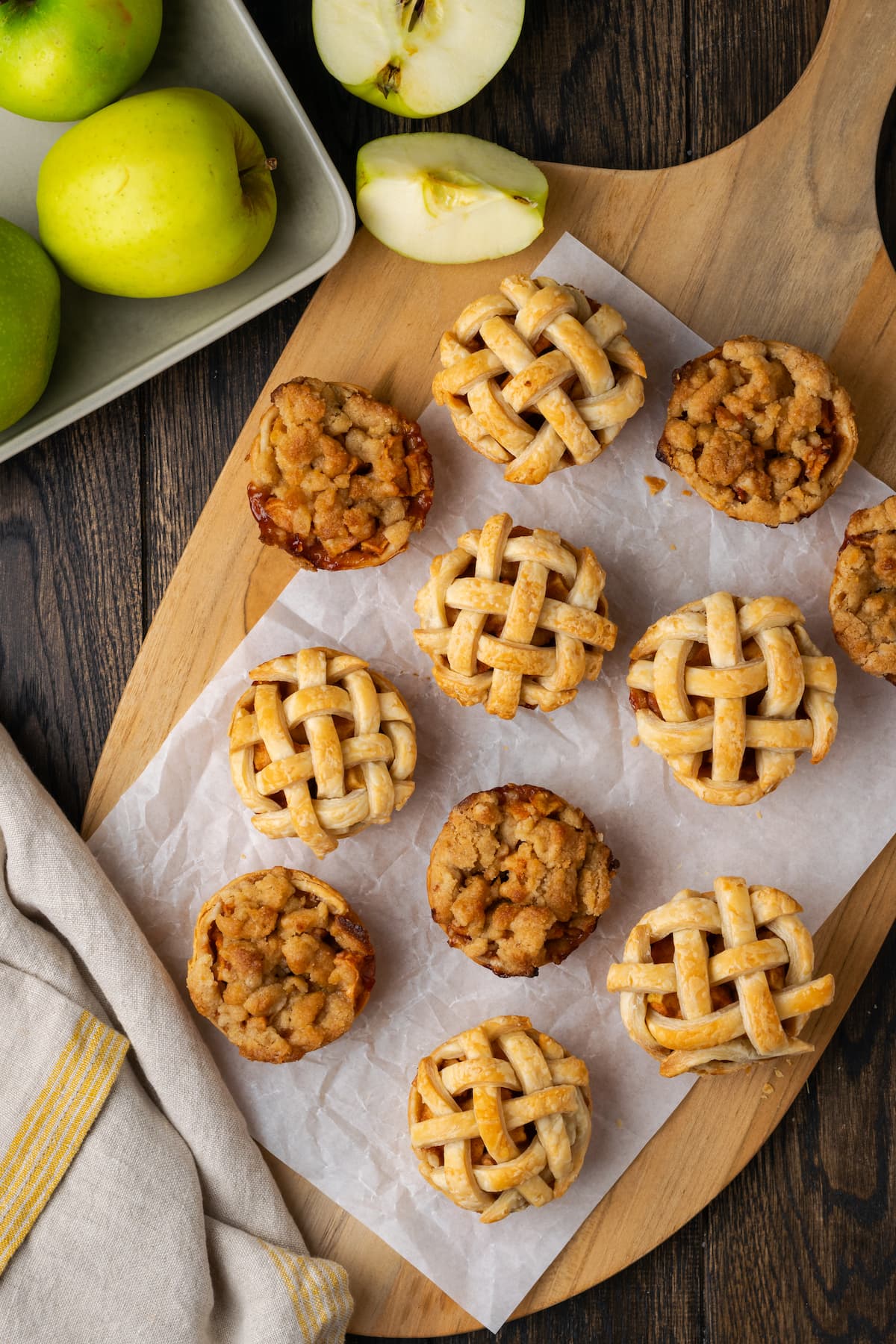 Mini apple pies are a bite-sized spin on a classic fall dessert! So easy to make and bursting with juicy apple pie filling and warm spices. Overhead view of mini apple pies on a wooden board lined with a piece of parchment paper, next to granny smith apples in a bowl.