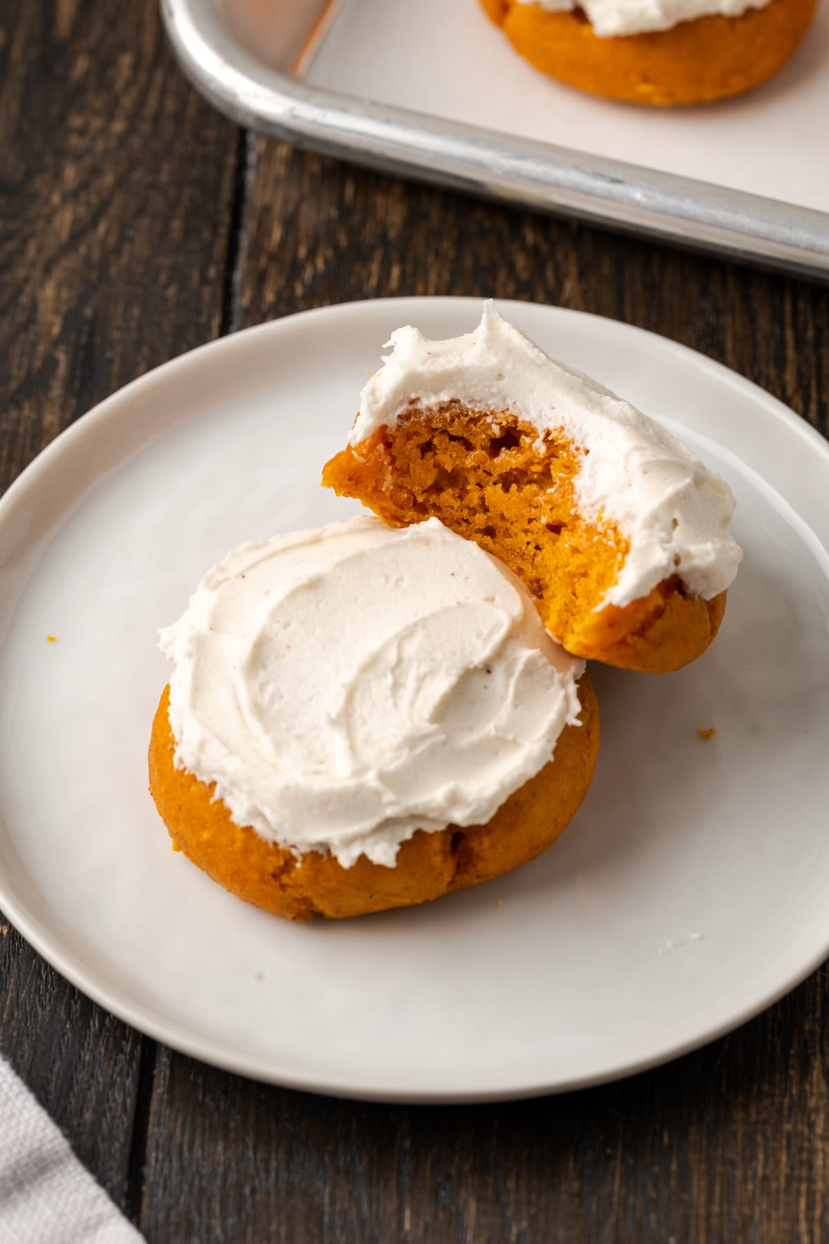 These soft and fluffy pumpkin cookies with cake mix are an easy fall dessert, made with only 2 ingredients and ready for the oven in minutes! Two frosted cake mix pumpkin cookies on a white plate, with a bite missing from one cookie.