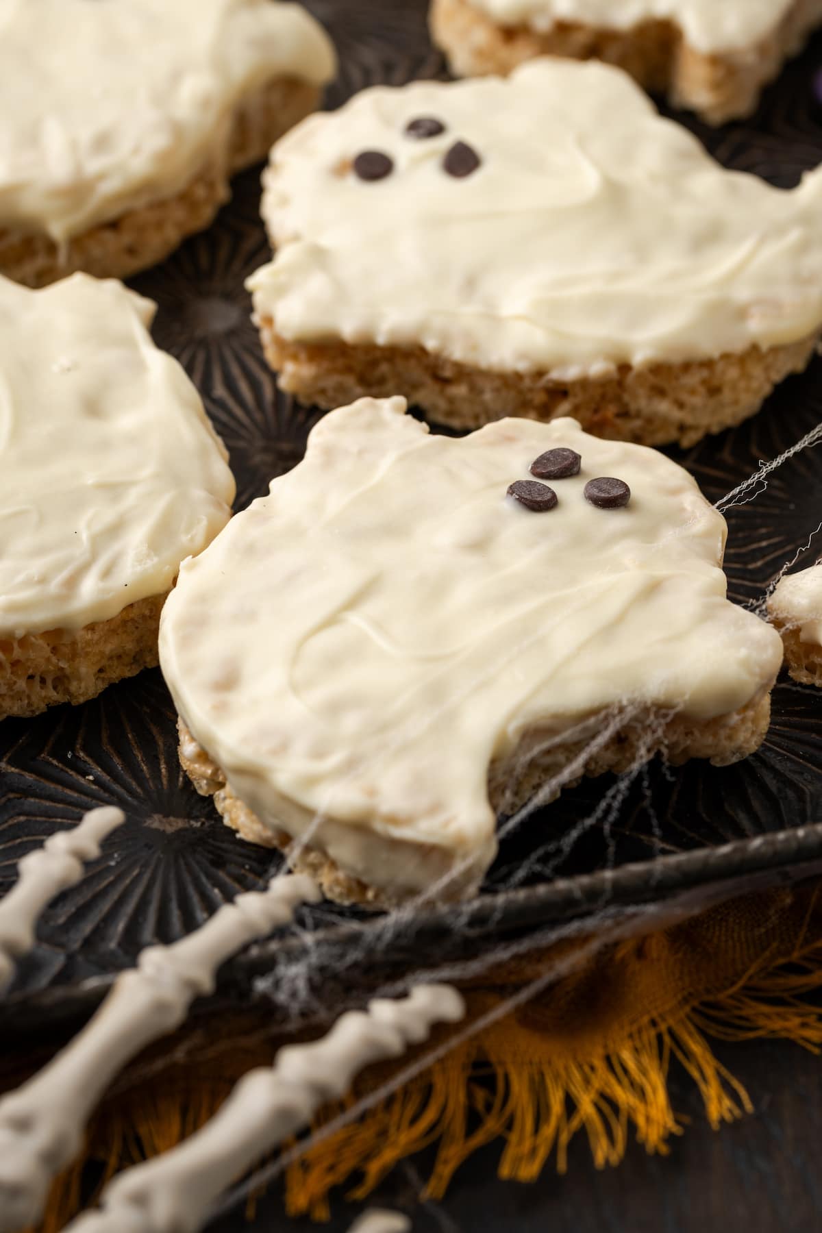 Halloween rice krispie treat ghosts on a baking sheet, decorated with faces made from mini chocolate chips, with a plastic skeleton's fingers in the corner of the frame.