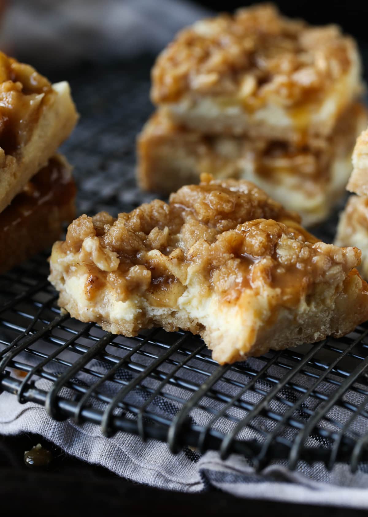 Caramel Apple Cheesecake Bars are creamy, sweet and delicious. They're a simple, buttery crust with an easy apple cheesecake filling topped with an oat streusel and caramel. A cheesecake bar on a wire rack with a bite taken out.